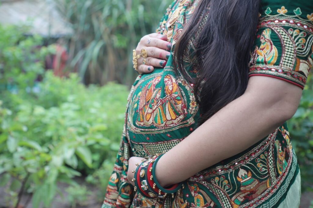 Pregnant woman in festive attire embracing maternity in lush garden setting, Dhari, Gujarat.